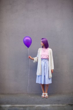 Gri duvarın önünde duran mor saçlı kız ve airballoon tutun