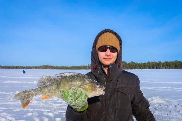 Young man is holding frozen fish after successful winter fishing at cold sunny day. Winter sport fishing fishing catch big bass — Stock Photo, Image