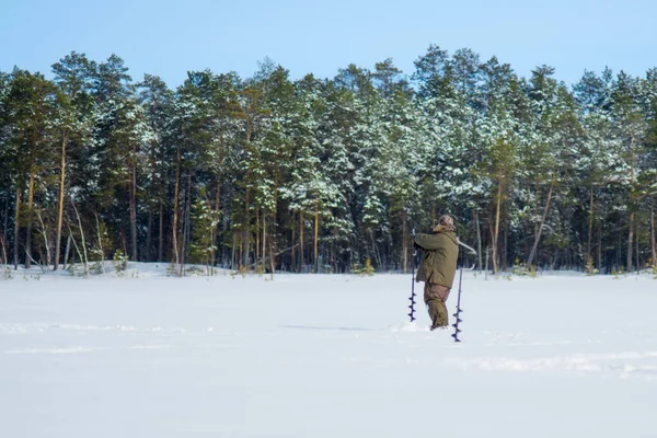 Man fisherman drills ice on the background of winter forest winter sport winter fishing — Stock Photo, Image