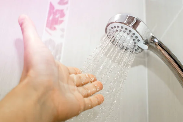 Mão masculina sob o fluxo de água da cabeça do chuveiro, fixado no suporte, verifica a temperatura da água no banheiro — Fotografia de Stock