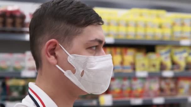 Молодой человек в маске в гипермаркете, покупает товары во время пандемии ковида 19. селективная фокусировка, движение камеры — стоковое видео