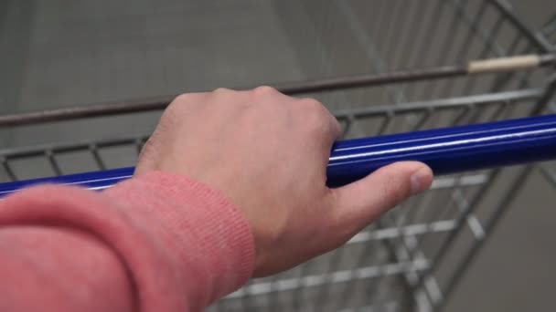 La mano masculina sostiene la manija de un carro de la compra rueda en la baldosa en la tienda. Vista en primera persona de cerca . — Vídeos de Stock