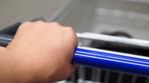 Mužská levá ruka drží držadlo nákupního košíku, tlačí poličku v supermarketu. Zavřít pohled první osoby.