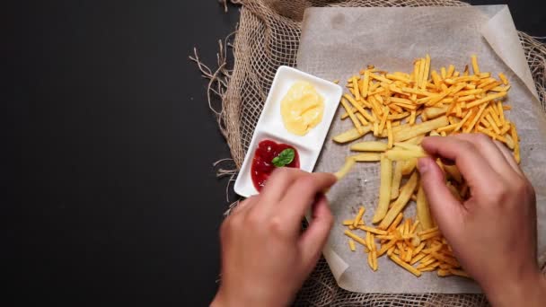 Homem come batatas fritas com ketchup e molho de queijo em uma mesa escura, vista superior. deitado plano — Vídeo de Stock