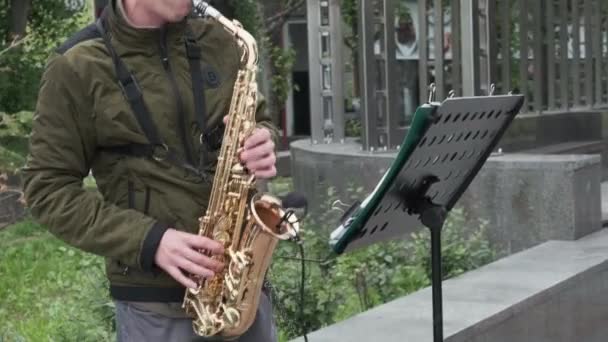 一个穿着绿色夹克和灰色裤子的街头音乐家在街上演奏铜萨克斯管 你看不到他的头 — 图库视频影像