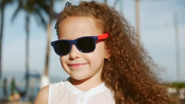 Крупный план портрета счастливой милой девочки с вьющимися волосами и красными солнечными очками, смотрящей в камеру — стоковое видео