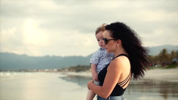 Família jovem, uma mãe carregando um filho em seus braços. Uma família feliz caminhando ao longo da costa . — Vídeo de Stock