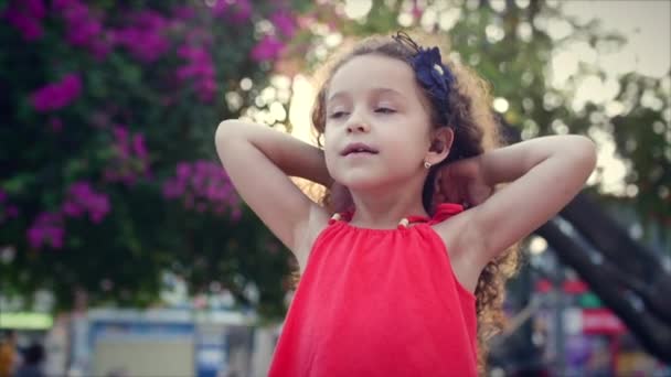 Beautihul menina Corrige o cabelo encaracolado. Retrato de menina bonita perto de árvore florescida no parque. Conceito de bebê e beleza natural . — Vídeo de Stock