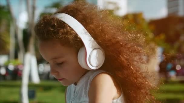 Kaukázusi aranyos lány göndör haj, fehér ruha ül a parkban, a fehér fejhallgató, zenét hallgatni, néz a távolba. — Stock videók