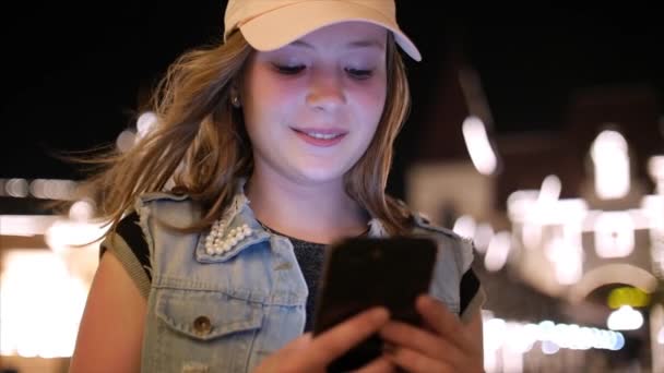 Atractiva adolescente usando un teléfono móvil mientras camina por las calles en una ciudad nocturna, en la ciudad nocturna de fondo. Material de archivo . — Vídeos de Stock