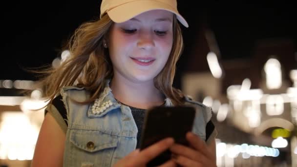 Atractiva adolescente usando un teléfono móvil mientras camina por las calles en una ciudad nocturna, en la ciudad nocturna de fondo. Material de archivo . — Vídeos de Stock