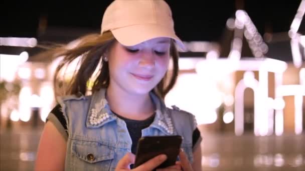 Atractiva adolescente usando un teléfono móvil mientras camina por las calles en una ciudad nocturna, en la ciudad nocturna de fondo. Material de archivo . — Vídeos de Stock