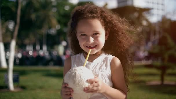 Bebé caucásico, niña con el pelo rizado, en un vestido blanco, beber coco . — Vídeos de Stock