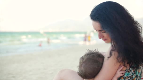 Mom with baby are enjoying fresh air sit by the sea, mom is breastfeeding the baby and lulling him. — Stock Video