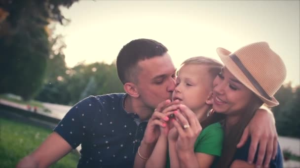 Gelukkige familie, blij blij vader, moeder en kind zitten op het gras, produceren sterke hugs, kus baby. — Stockvideo