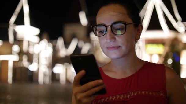 Chica atractiva usando un teléfono móvil mientras camina por las calles en una ciudad nocturna, en la ciudad nocturna de fondo. Material de archivo . — Vídeos de Stock
