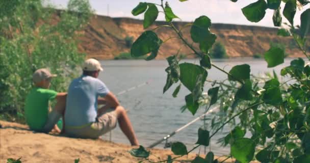 Un padre y un hijo felices están sentados en la orilla del río, la pesca, papá e hijo están pescando en la mañana. Familia feliz . — Vídeos de Stock