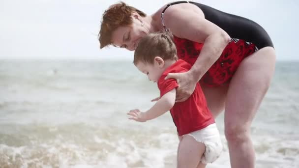 Un enfant heureux avec une grand-mère joue à la mer, le petit garçon sourit joyeusement, rit, saute sur les vagues, court le long du littoral. Concept de famille heureuse et amicale . — Video