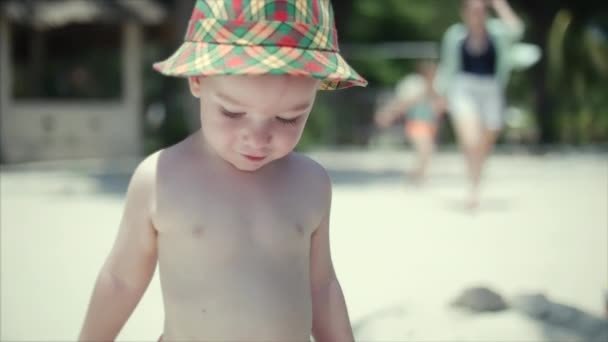 Fröhlicher Junge mit Hut geht am Sand entlang, im Hintergrund kommt seine Familie — Stockvideo