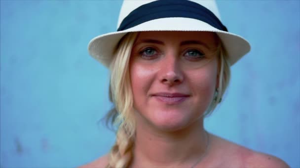 Retrato de uma aparência bonita mulher caucasiana, loira em um chapéu branco com fita azul, ao ar livre, sorrindo olhando para a câmera. Vista cortada, fundo azul . — Vídeo de Stock