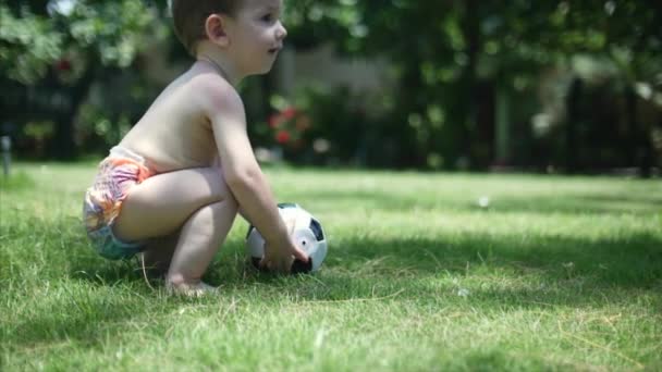 Um menino de dois anos gosta de jogar futebol no parque, correr e chutar a bola . — Vídeo de Stock