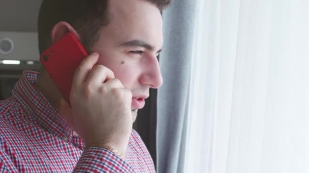 Närbild av ung man i en rutig skjorta talar på en röd telefon medan du tittar ut genom fönstret. 4 k. lager fotage — Stockvideo