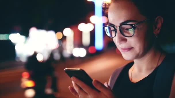 Attractive woman using a mobile phone while walking through the streets in a night city, in the background can see bikers. Stock footage. — Stockvideo