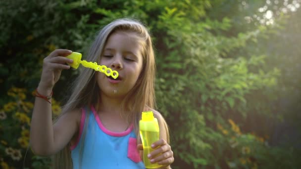 Slow motion av en glad liten kaukasiska flicka blåser såpbubblor i en solig dag. Begreppet lycklig barndom eller barnens spel i naturen. — Stockvideo