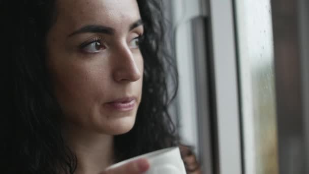 Mujer caucásica joven pensativa mirando la lluvia fuera de la ventana bebiendo café . — Vídeos de Stock