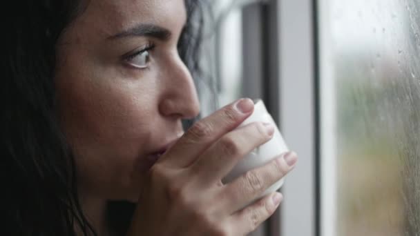 Mujer caucásica joven pensativa mirando la lluvia fuera de la ventana bebiendo café . — Vídeos de Stock