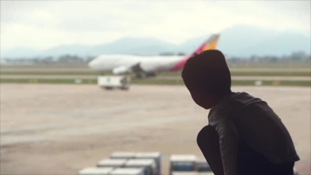 Airport. Little boy looks out the window with view on airport area, he watches as the plane leaves for the take-off strip. — Stock Video