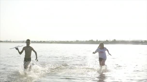 Infancia feliz y despreocupada. Los niños juegan con un perro, corren a lo largo de la arena, ríen, juegan en el río, lanzan un avión de juguete . — Vídeos de Stock