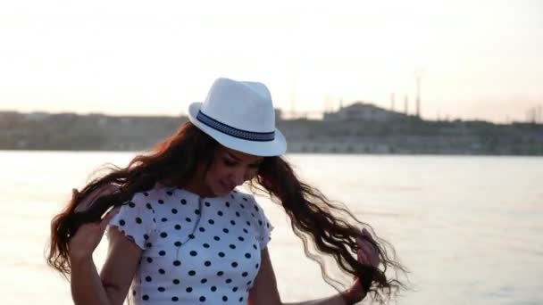 Menina feliz em um chapéu bonito branco caminha ao longo do litoral, na areia, olha para o pôr do sol, sorrindo — Vídeo de Stock