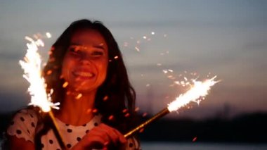 Gün batımında sahilde günbatımı, fireworks ile ağır çekimde maytap ile dans genç mutlu gülümseyen kadın.