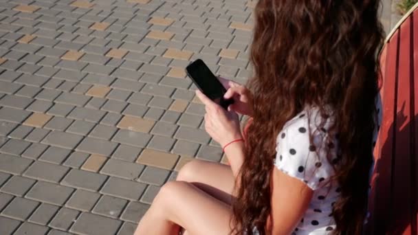 Mulher adolescente bonita de uma aparência caucasiana usando aplicativo de tecnologia de telefone inteligente sentado em um banco de ruas da cidade vivendo estilo de vida feliz urbano . — Vídeo de Stock