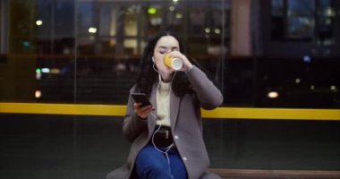 Gece city adlı arka plan sahip ile smartphone, kahve içme bir otobüs istasyonunda oturan güzel kadın.