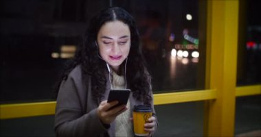 Gece city adlı arka plan sahip ile smartphone, kahve içme bir otobüs istasyonunda oturan güzel kadın.