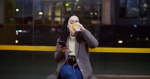 Mulher bonita sentada em uma estação de ônibus, bebendo café com smartphone, tendo cidade noturna ao fundo . — Vídeo de Stock