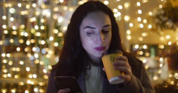 Mujer atractiva usando el teléfono móvil mientras camina por las calles en el fondo de las luces festivas de la ciudad nocturna . — Vídeo de stock