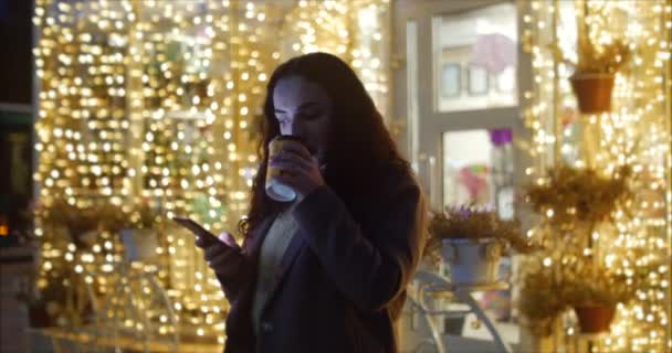 Atractiva mujer tomando café usando un teléfono móvil mientras camina por las calles en el fondo de las luces festivas de la ciudad nocturna. — Vídeo de stock