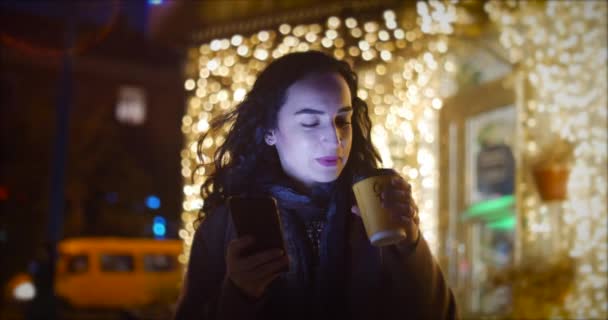 Mujer atractiva usando el teléfono móvil mientras camina por las calles en el fondo de las luces festivas de la ciudad nocturna . — Vídeos de Stock