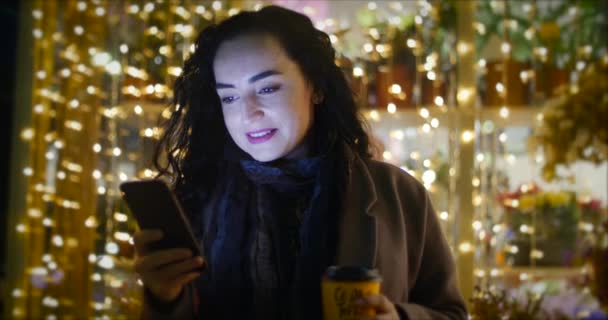 Atractiva mujer tomando café usando un teléfono móvil mientras camina por las calles en el fondo de las luces festivas de la ciudad nocturna. — Vídeos de Stock