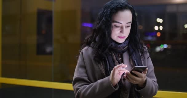 Attractive woman using mobile phone while walking through the streets on the background of festive lights of night city. — Stock Video