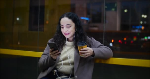 Vacker kvinna sitter på en busstation, dricka kaffe med smartphone, med natt staden i bakgrunden. — Stockvideo