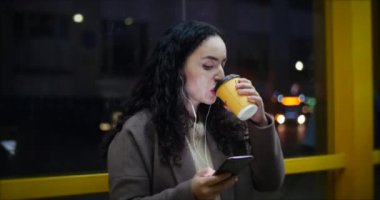 Gece city adlı arka plan sahip ile smartphone, kahve içme bir otobüs istasyonunda oturan güzel kadın