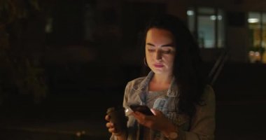 Güzel moda mutlu şık genç kadın Jean ceket beyaz kulaklık ve uzun karanlık kıvırcık saç meşgul ile onun cep telefonu iken yürüme ile bir şehir sokak. Güzel kız üzerinde akıllı ileti