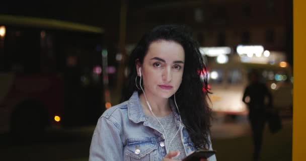 Hermosa mujer joven con estilo feliz de moda en chaqueta Jean con auriculares blancos y largo pelo rizado oscuro ocupado con su teléfono móvil mientras camina por una calle de la ciudad. Mensajería de chica bonita en el Smart — Vídeos de Stock