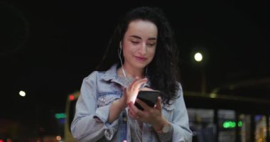 Güzel moda mutlu şık genç kadın Jean ceket beyaz kulaklık ve uzun karanlık kıvırcık saç meşgul ile onun cep telefonu iken yürüme ile bir şehir sokak. Güzel kız üzerinde akıllı ileti