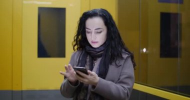 Güzel moda mutlu şık genç kadın kahverengi ceket koyu mavi eşarp ve uzun karanlık kıvırcık saç sahiptir kahve meşgul ile onun cep telefonu iken yürüme ile bir şehir sokak. Güzel kız mesajlaşma