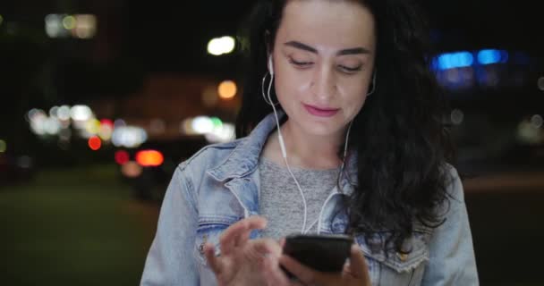Beautiful Fashionable Happy Stylish Young Woman in Jean Jacket with White Headphones and Long Dark Curly Hair Busy with her Mobile Phone While Walking a City Street. Pretty Girl Messaging on the Smart — Stock Video